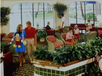 a  couple walking through a seating area. A woman dressed in a blue dress is holding a mickey mouse plushey. A blonde man in a red shirt and chinos is standing next to her. They are the focus of the image